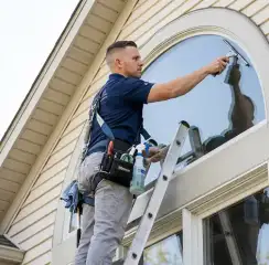 Professional window cleaner on a ladder using a squeegee to clean an exterior arched window, Jacksonville, FL.